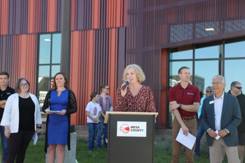 Woman standing outside at lectern holding a mic to her mouth. 