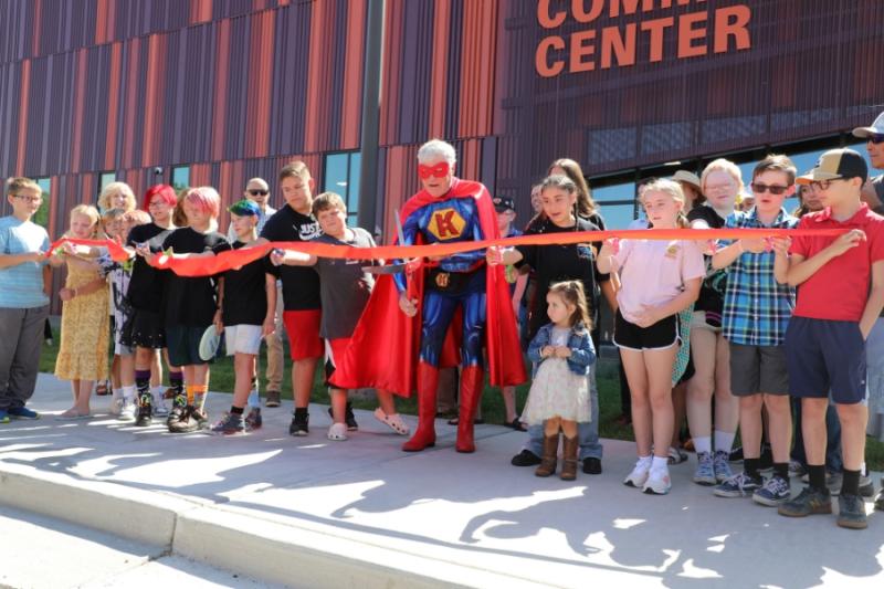 Large group of people standing around man dressed as superhero holding large red scissors. 