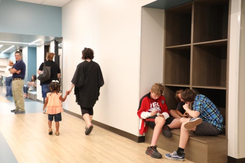 Woman holding toddler's hand walking in hall next to three kids sitting on bench. 