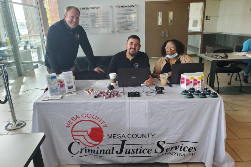 Two people sitting and one standing at a white table with black text reading, "Mesa County Criminal Justice Services Department," with the red Mesa County logo.