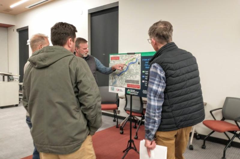 Three people facing a man pointing at a poster board with a map on it. 