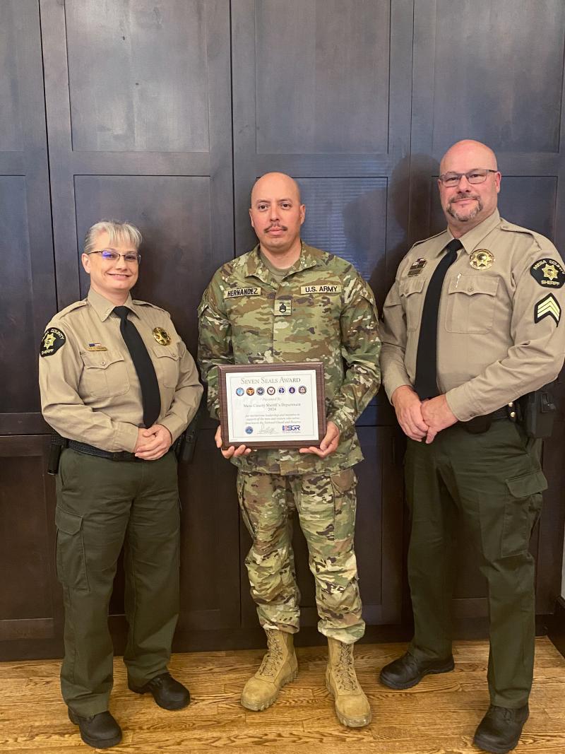 Deputy Barry Hernandez (a tall bald male in an National Guard uniform) stands between Lieutenant Peck (left - a short woman with blond hair pulled into a bun wearing glasses and a tan uniform) and Sergeant Craig Bowen (right - a tall bald male with glasses in a tan uniform) holding the Seven Seals Award.