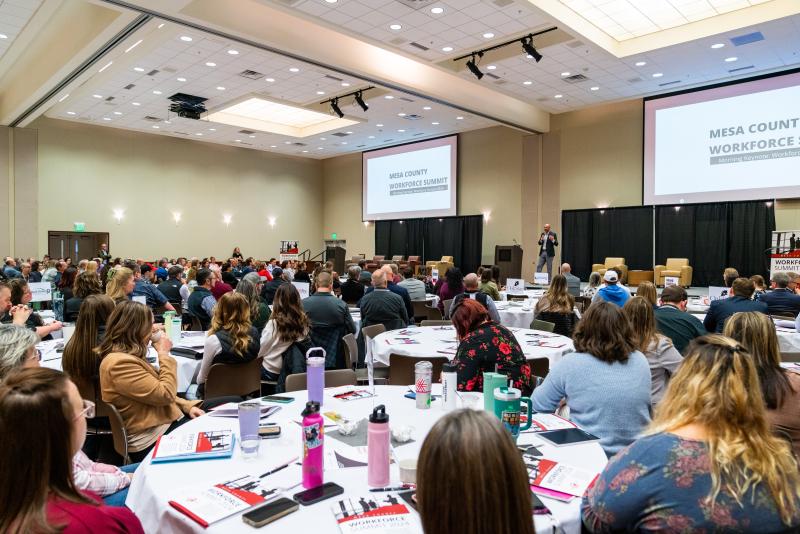 Presenter talking to a full conference room of engaged people sitting around tables. 