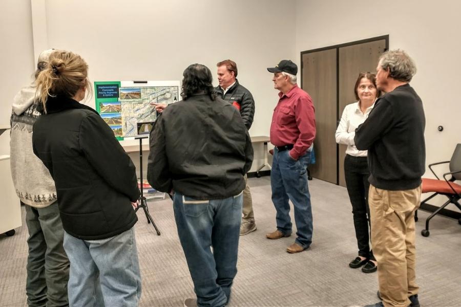 Three people facing a man pointing at a poster board with a map on it. 