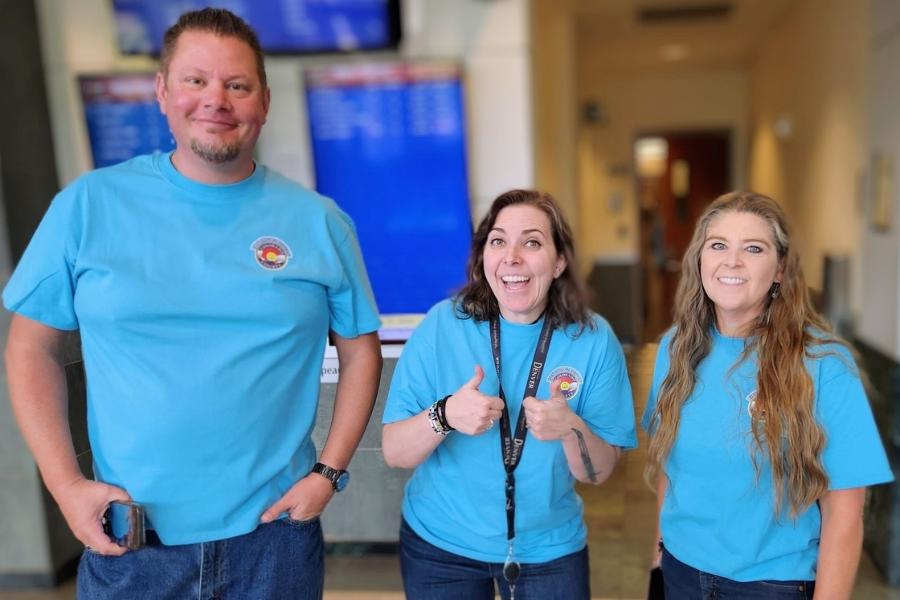 Three people wearing blue shirts and smiling at camera. The middle individual is giving two thumbs up.