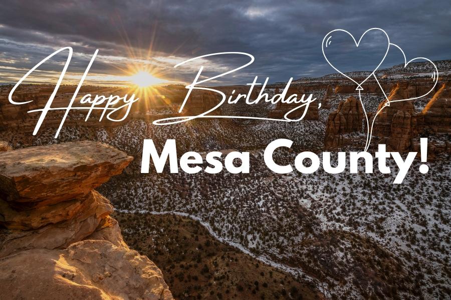 Snow covered dark green bushes under huge red rocks with white text reading, Happy Birthday, Mesa County. 