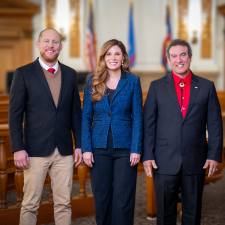 A group photo of three commissioners standing in an official government setting. They are dressed in formal attire, with an American flag and other banners in the background.