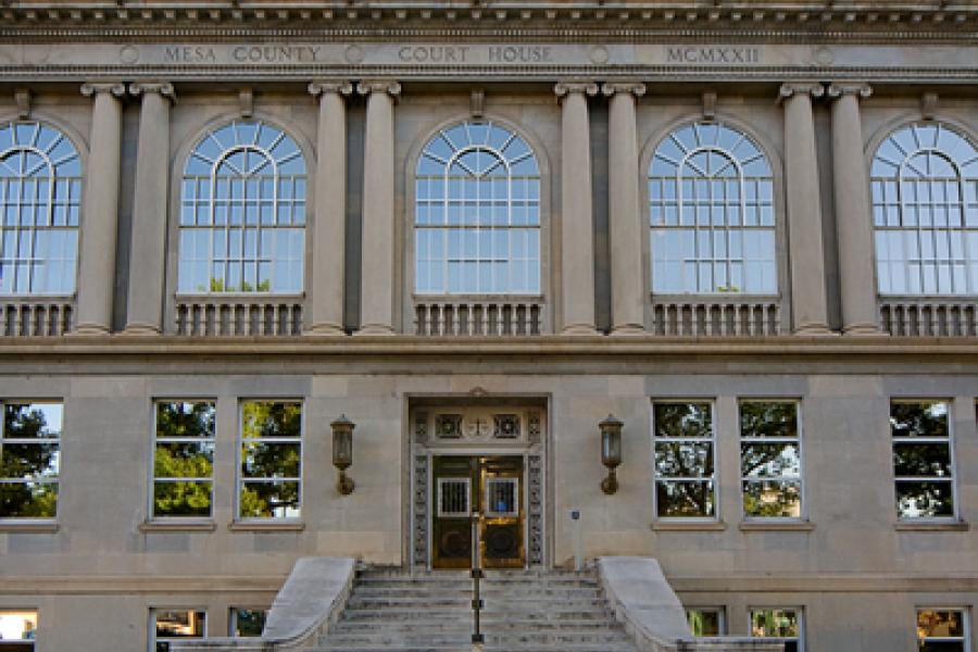 Photograph of the front of old Mesa County Courthouse showing the wide staircase, four park benches, trees, flowering plants, shrubs, and grass area