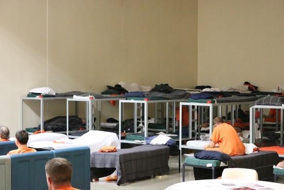 Historical photograph of a group jail cell with bunk beds, beds, chair, and inmates in orange uniforms