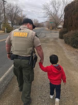 Sherriff walking in uniform holding the hand of a young boy in a red sweatshirt.