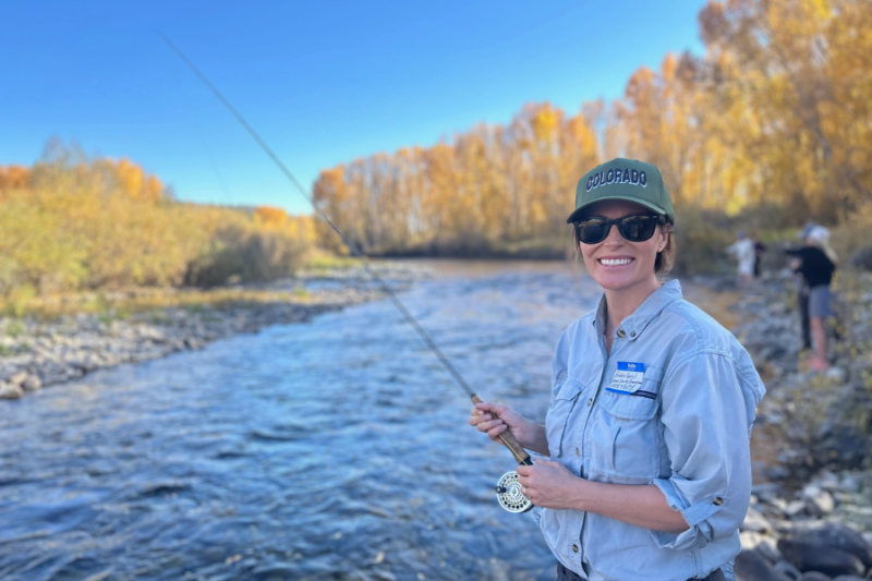 A woman, identified as Bobbie Daniel by her name tag, stands by a river holding a fishing rod. She is wearing a green "Colorado" cap, sunglasses, and a light blue shirt. The trees in the background are golden, indicating autumn