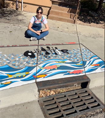 Artist sitting next to her stormwater drain art that features a fish painted to represent trash.