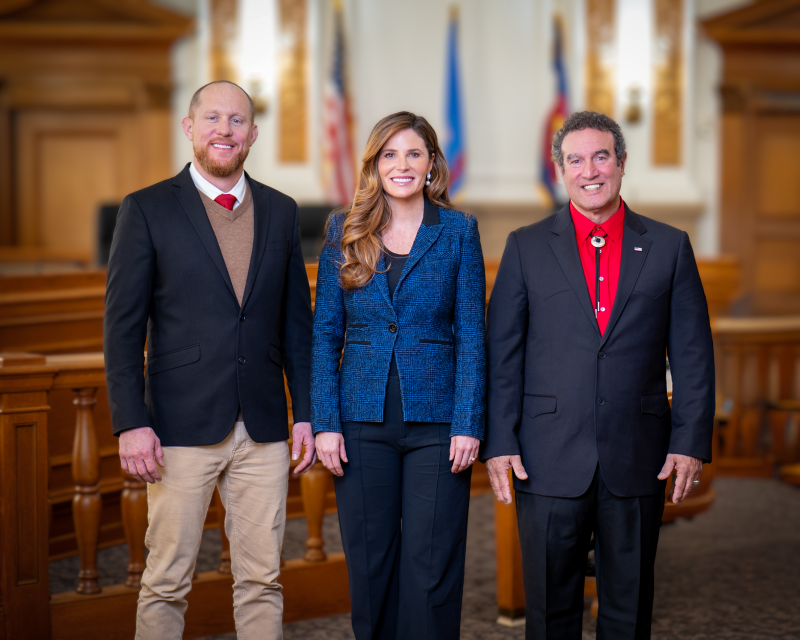 A group photo of three commissioners standing in an official government setting. They are dressed in formal attire, with an American flag and other banners in the background.