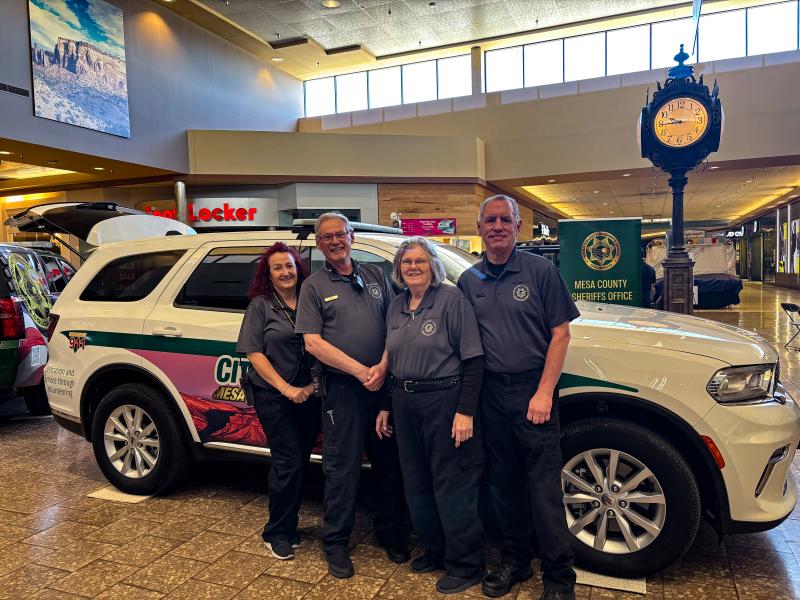 A group of volunteers wearing matching grey Mesa County Sheriff's Office Volunteer polo shirts and black pants stand against a Citizens on Patrol vehicle. From left to right short female with red hair, taller bald male, short female with grey hair, tall male with short grey hair.