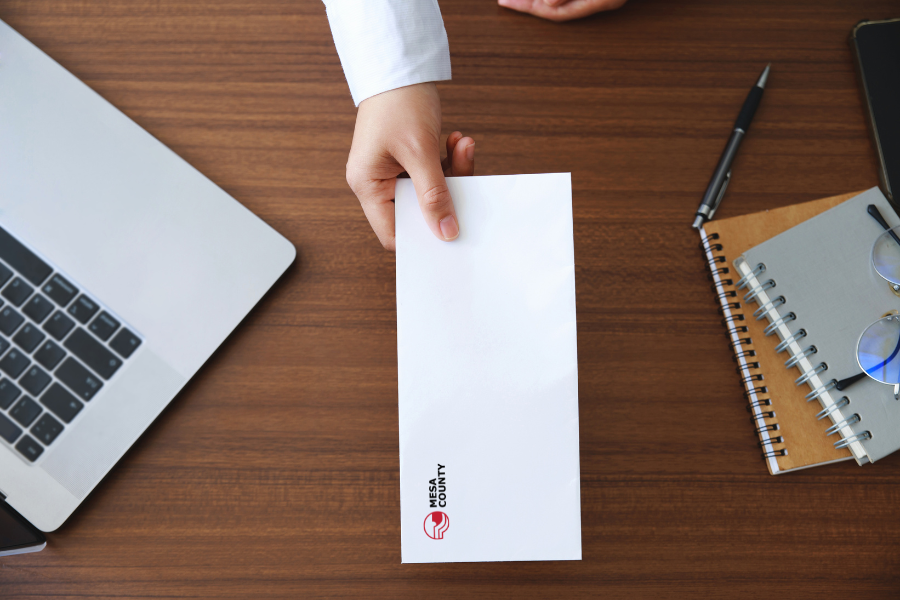 Hand holding white envelope with Mesa County logo on it. 