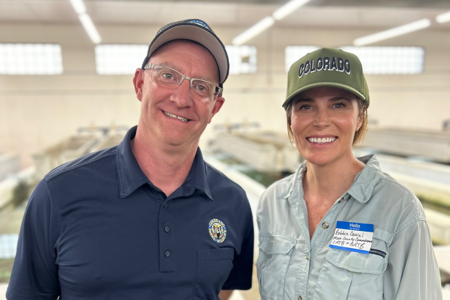 Man and woman smiling at a fish hatchery, wearing caps and casual outdoor attire, with the woman identified as Bobbie Daniel from Mesa County.