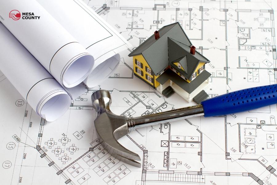 White building plans with tiny house and hammer laid on top of the plans with a red Mesa County logo on the top left corner. 