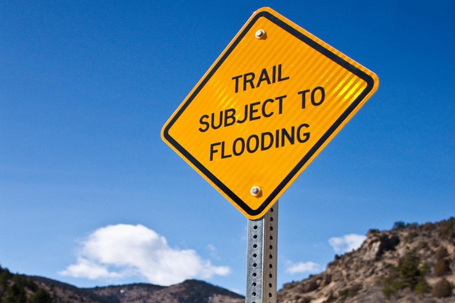 A yellow sign that reads "TRAIL SUBJECT TO FLOODING" in all caps black text. A rocky canyon with a river running through it sits below a blue sky in the background.