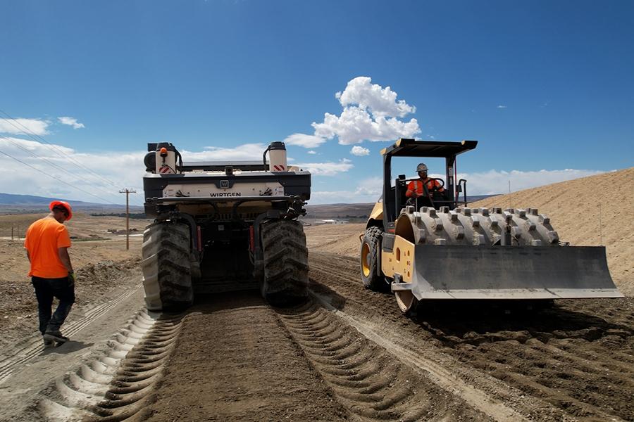 Two pieces of heavy equipment working on a road project with two construction workers present. 