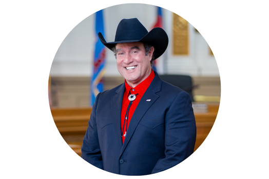 Headshot-style portrait of a smiling person in a dark suit with a red shirt, wearing a dark cowboy hat.