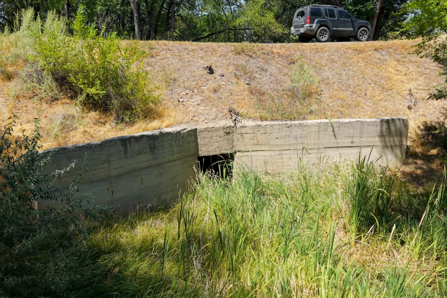 Photo of a drainage structure on 36 Road. The culvert is in a dip in the roadway, surrounded by brush and trees.