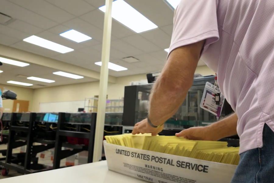 An election official prepares mail ballots for processing through a ballot counting machine. The dozens of yellow ballots are in a tray on a white table in the elections building. 